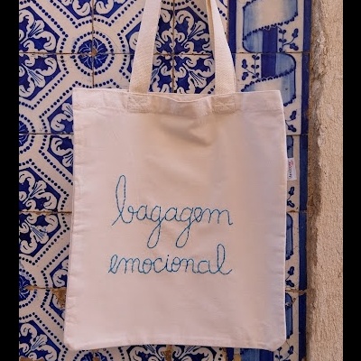 Saco branco com texto 'bagagem emocional' pendurado em parede de azulejos azuis e brancos
