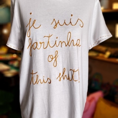 T-shirt branca com texto bordado castanho 'je suis fartinha of this shit' num manequim preto em loja