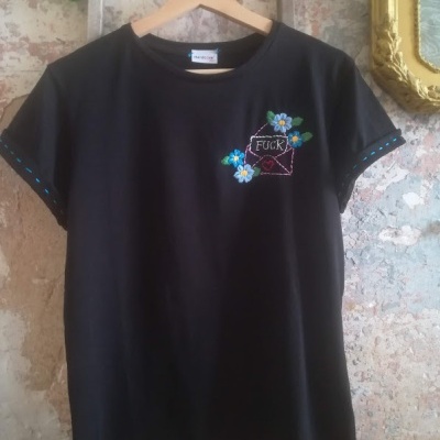T-shirt preta com bordado floral colorido e texto 'FUCK', pendurada em cabide de madeira