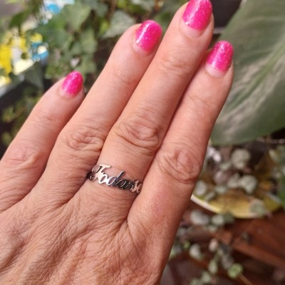 Mão feminina com anel prateado e unhas cor-de-rosa brilhantes, várias plantas ao fundo