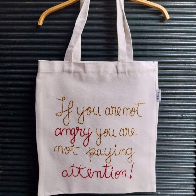 Saco de tecido bege com texto bordado e alças pendurado num cabide