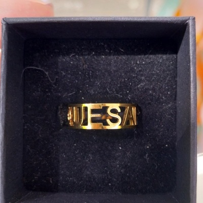 Anel dourado com letras vazadas em caixa preta.