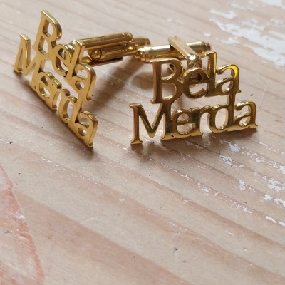 Botões de punho dourados com texto Bela Merda sobre madeira clara