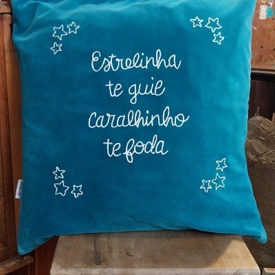 Almofada azul de veludo com bordado branco e texto em português sobre cadeira de madeira