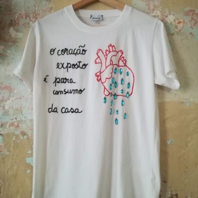 T-shirt branca com bordado de coração vermelho e gotas azuis, texto bordado em preto, pendurada numa cabide de madeira