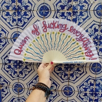 Leque branco decorado com frase em vermelho, segurado à frente de azulejos azuis e brancos.