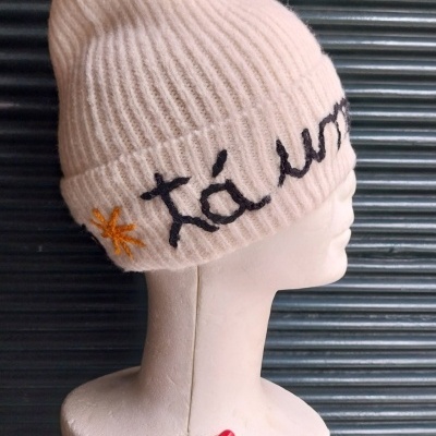 Gorro de malha bege claro bordado com texto em preto e amarelo numa cabeça de manequim branca.
