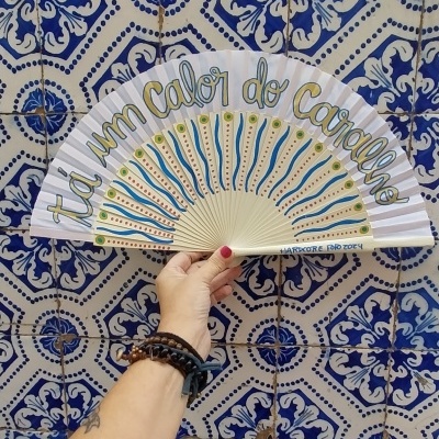 Leque branco com desenhos coloridos e texto, segurado à frente de azulejos azuis e brancos decorativos.