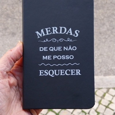 Caderno preto com texto branco segurado por mão, calçada portuguesa ao fundo