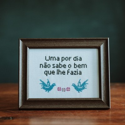 Placa decorativa bordada, moldura de madeira castanha, frase em português, pássaros azuis e flores rosas