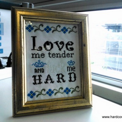 Quadro bordado com texto 'LOVE me tender AND me HARD' em moldura dourada junto a janela