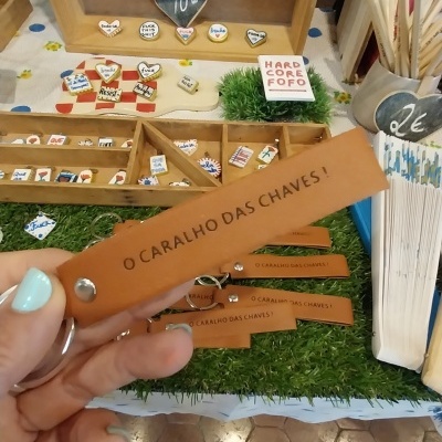 Chaveiro em pele castanha com texto O CARALHO DAS CHAVES! e exposição de outros produtos em madeira e tecido