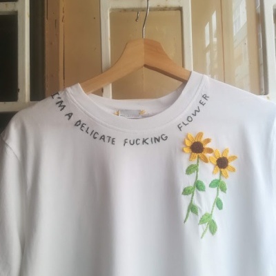 T-shirt branca com flores bordadas amarelas e texto preto na gola pendurada num cabide de madeira