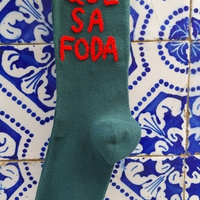 Meia verde escura com texto vermelho 'QUE SA FODA' sobre azulejos azuis e brancos