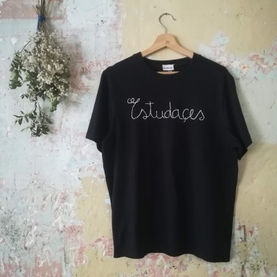 T-shirt preta com texto 'Estudages' bordado branco pendurada na parede com flores ao lado