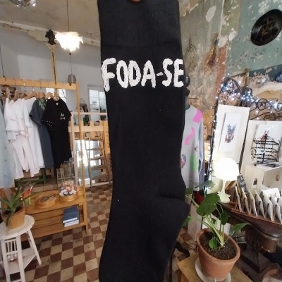 Meia preta com a palavra FODA-SE escrita em branco, segurada à mão no interior de loja com roupas e plantas.