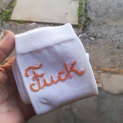 Meia branca com bordado laranja da palavra 'Fuck' segurada à mão