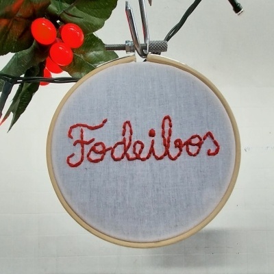 Bastidor de bordado com a palavra 'Fodeiros' em linha vermelha e elementos decorativos verdes com bagas vermelhas.