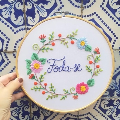 Bordado colorido em aro de madeira com palavra 'Foda-se' e flores, contra azulejos azuis e brancos.