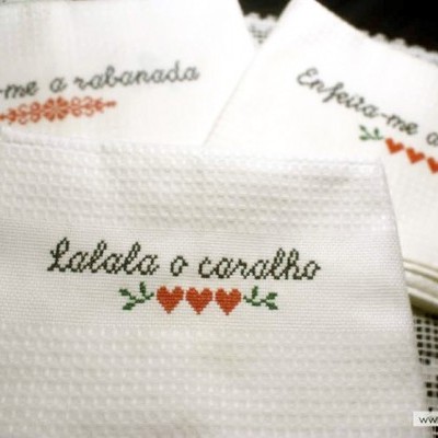 Três toalhas brancas com bordados em ponto cruz com frases em português e corações coloridos.