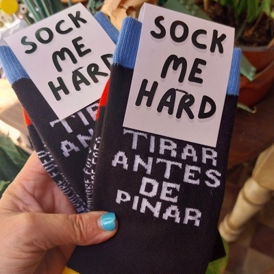 Par de meias pretas com punho azul e ponta amarela, texto nas meias e etiquetas, seguidas em ambiente com plantas