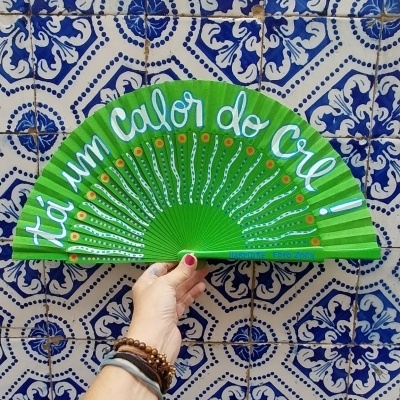 Leque verde com texto branco 'tá um Calor do Cru!' e fundo de azulejos azuis e brancos.