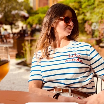 Blusa branca com riscas azuis e texto bordado vermelho usada por mulher com óculos de sol num café ao ar livre
