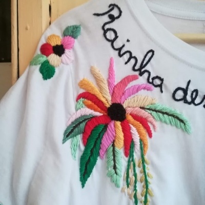 T-shirt branca com bordados florais coloridos e texto 'rainha de' em preto
