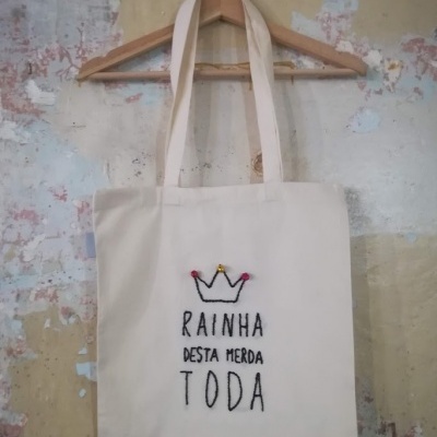 Saco de tecido cru com bordado colorido e texto preto pendurado num cabide