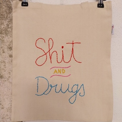 Pano de algodão bege com bordado colorido a dizer 'Shit AND Drugs'