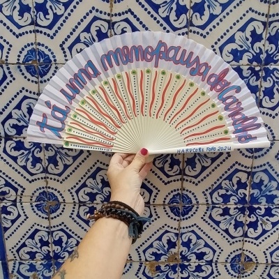 Leque branco pintado com texto e padrões coloridos contra parede de azulejos azuis e brancos.