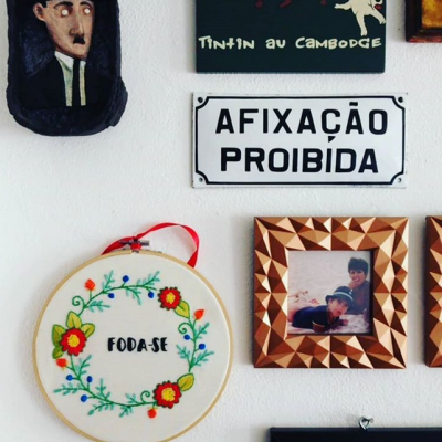 Parede branca com quadros decorativos incluindo placa 'AFIXAÇÃO PROIBIDA' e bordado com palavra 'FODA-SE'.