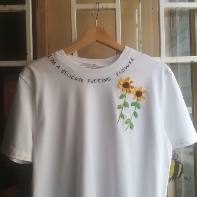 T-shirt branca com bordado de flores amarelas e texto no colarinho pendurada num cabide de madeira