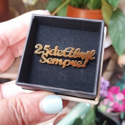 Peça de joalharia dourada com a frase 25 de Abril Sempre numa caixa preta