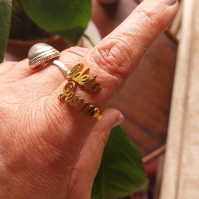 Dois anéis, um prateado com pérola e outro dourado com texto, no dedo de uma mão com plantas ao fundo.