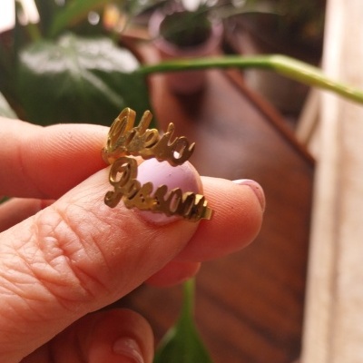 Anel dourado com inscrições Liberta Pessoa segurado por mão com unhas pintadas