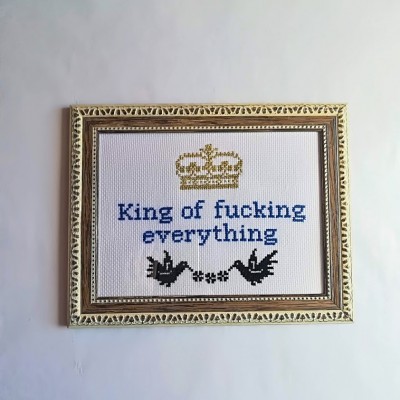 Quadro bordado com moldura dourada, coroa amarela, texto azul e pássaros pretos