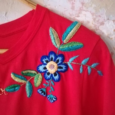 Camisola vermelha com bordado floral azul e verde, pendurada em cabide junto a parede desgastada