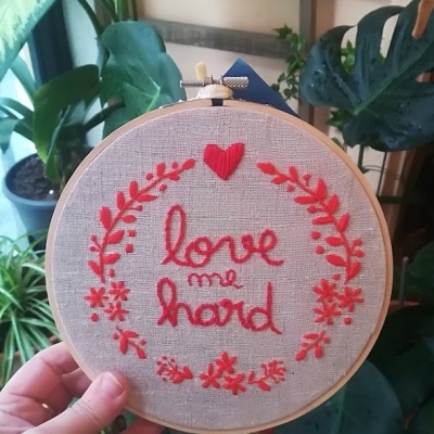 Aro de bordado com tecido e texto bordado em vermelho segurado por uma mão
