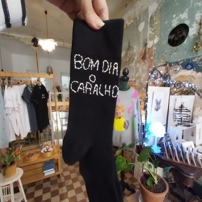 Meia preta com texto bordado branco 'BOM DIA O CARALHO' na mão em loja de roupa rústica