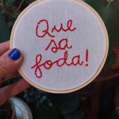 Bastidor de bordado com tecido branco e texto vermelho segurado por mão com unhas azuis