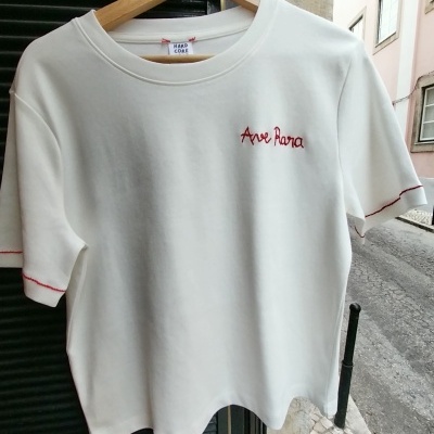 T-shirt branca com bordado vermelho Ave Rara e detalhes vermelhos nas mangas pendurada em cabide de madeira