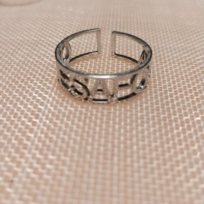 Anel prateado aberto com palavra recortada SAHCO sobre superfície bege texturada