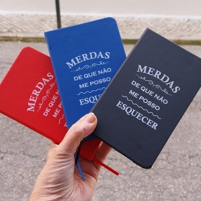 Capas de cadernos em vermelho, azul e preto com texto impresso em branco seguradas por mão com pulseiras