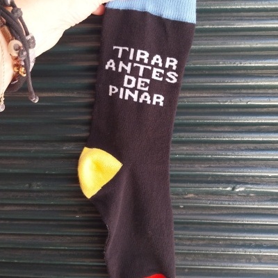 Meia preta com punho azul claro, calcanhar amarelo, ponta vermelha, texto branco, sendo segurada por mão com unhas azuis