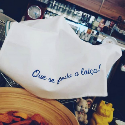 Pano de cozinha branco com texto azul bordado pendurado sobre escorredor metálico, utensílios de cozinha ao fundo.