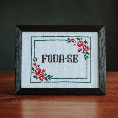 Quadro ponto cruz com texto FODA-SE e flores em moldura preta sobre mesa de madeira