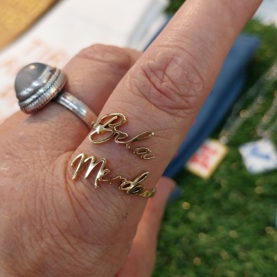 Anel dourado com texto Bela Merda num dedo de mão com anel prateado de pedra cinza e fundo verde desfocado