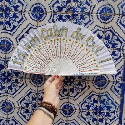 Leque branco com texto 'TÁ um Calor do CRA!' segurado diante de azulejos portugueses azuis e brancos