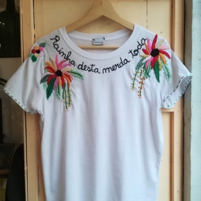 T-shirt branca com bordados coloridos e texto 'Rainha desta merda toda' no decote